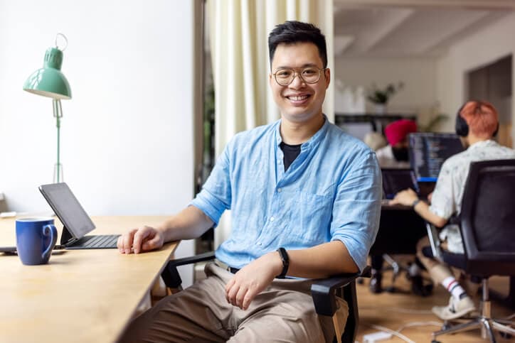 Korean man working looking at camera smiling while sitting at a coworking office.
