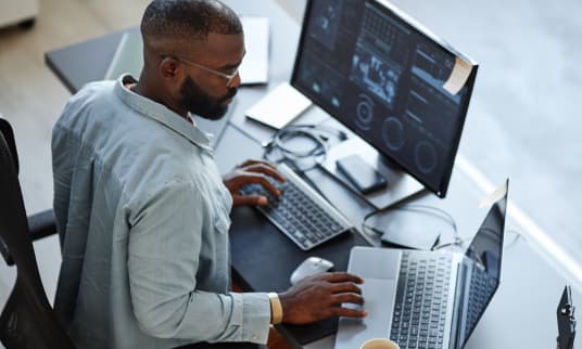 Man on desktop computer typing while looking at a laptop screen to his side.