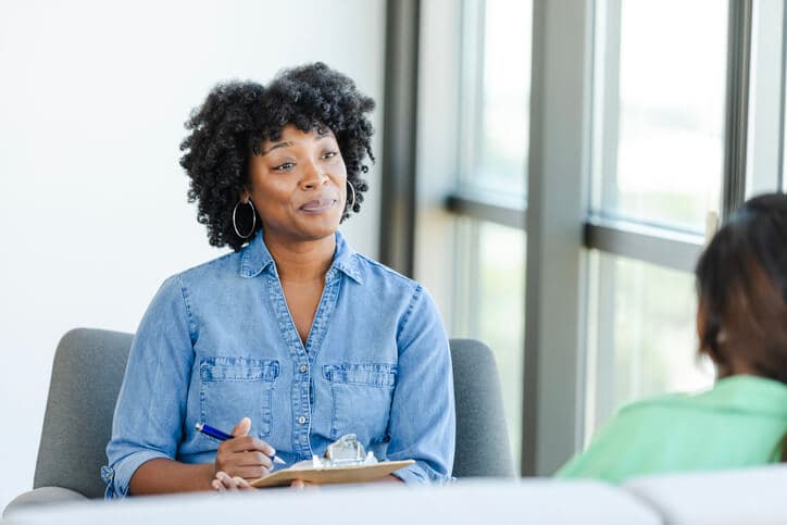 A mid adult Black female social worker takes notes while listening to a client during a counseling session.