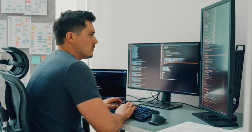 Male software developer sitting at a desk with multiple desktop screens at an office. He is coding an application program for AI technology development.