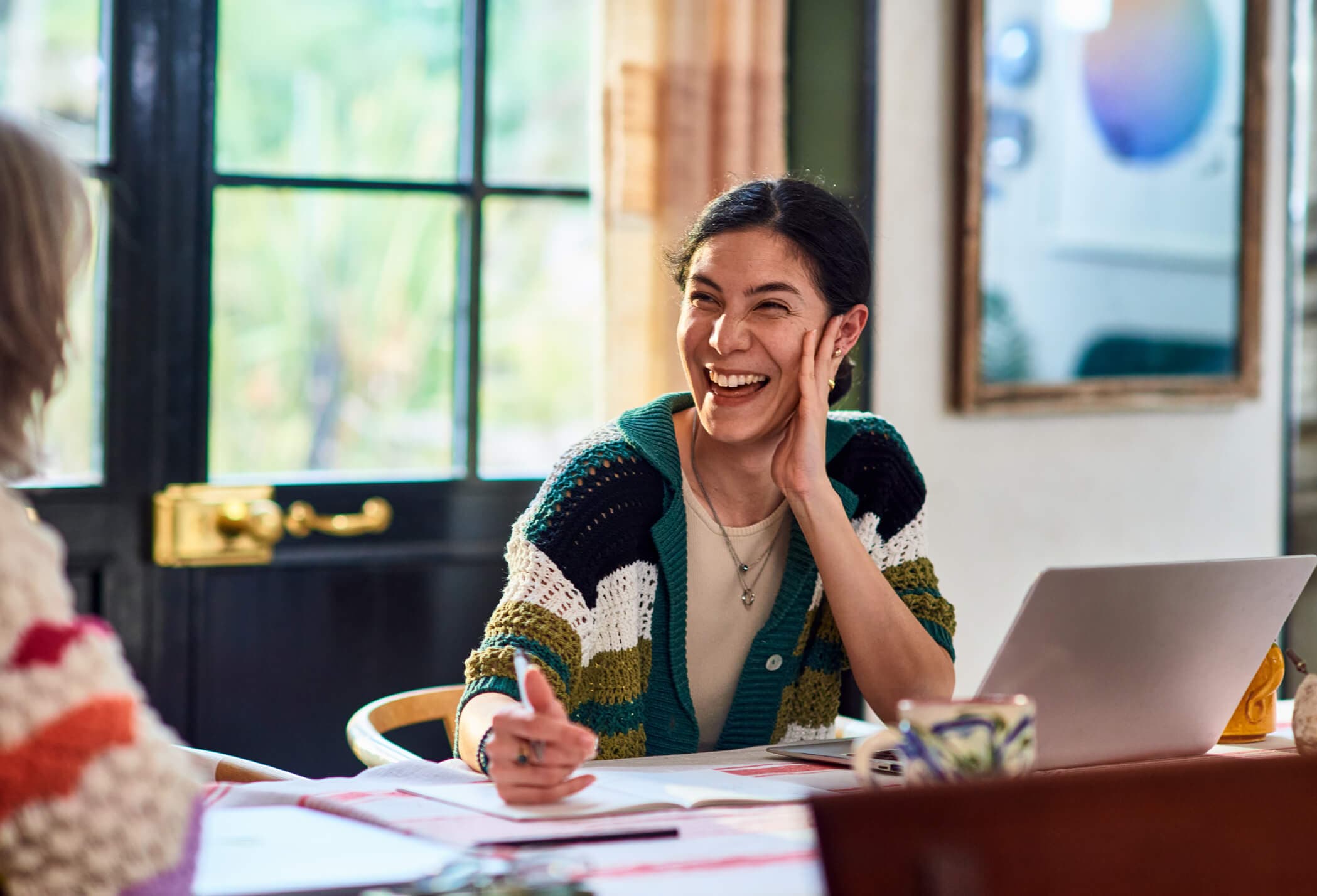 Smiling woman in her 30s sitting at a table talking and laughing with her colleague.