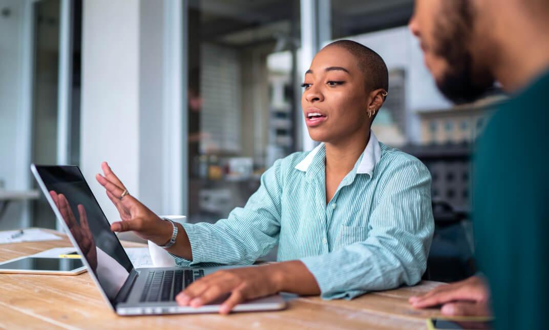 Black female business professional sits at an office meeting room table next to a male colleague. She is describing a set of data reports on her laptop machine.