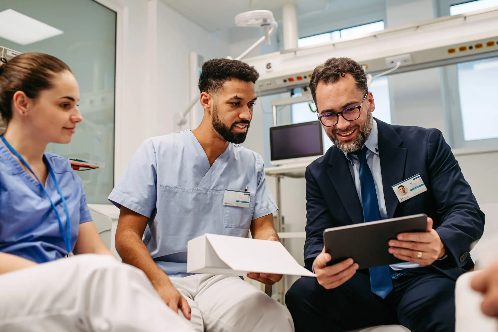 Hospital healthcare manager sharing info on a tablet device to two nursing team members in a hospital meeting room.