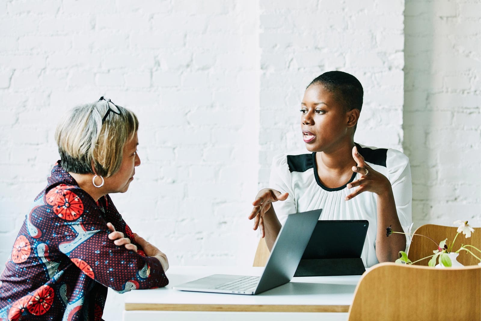 Two women engaged in conversation, each with a laptop open in front of them.